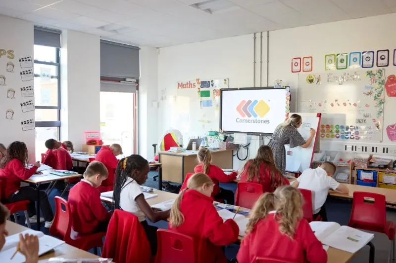 Primary school pupils working at desks while a teacher writes on a flipchart
